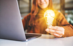 Woman with a laptop holding a lightbulb that is "lit up" by her idea.