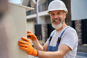 Waist-up portrait of cheerful electrical engineer in safety helmet installing external power distribution box